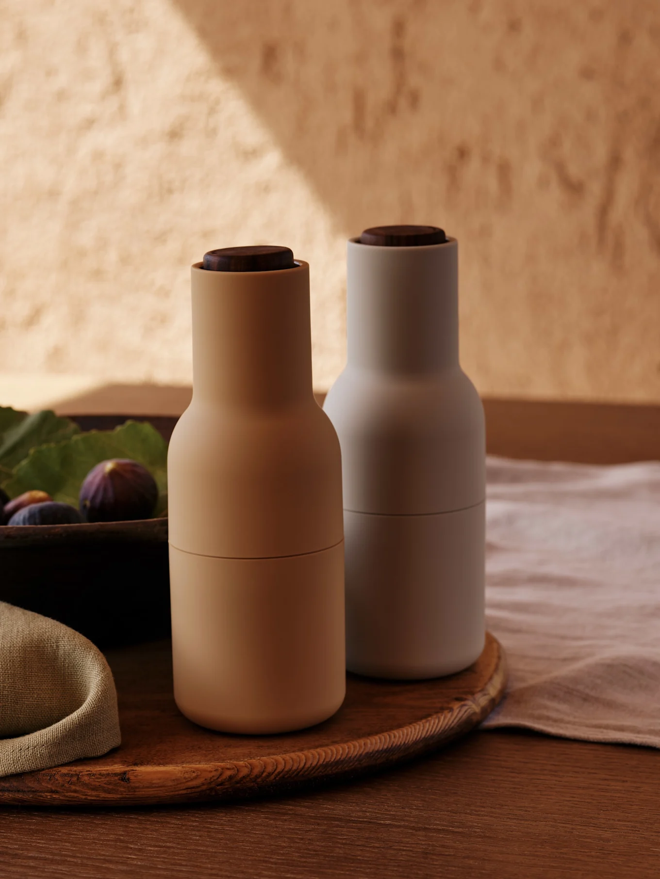 To moderne krydderkverner med tretopper, en beige og en lys grå, på et rundt trefat med fiken.