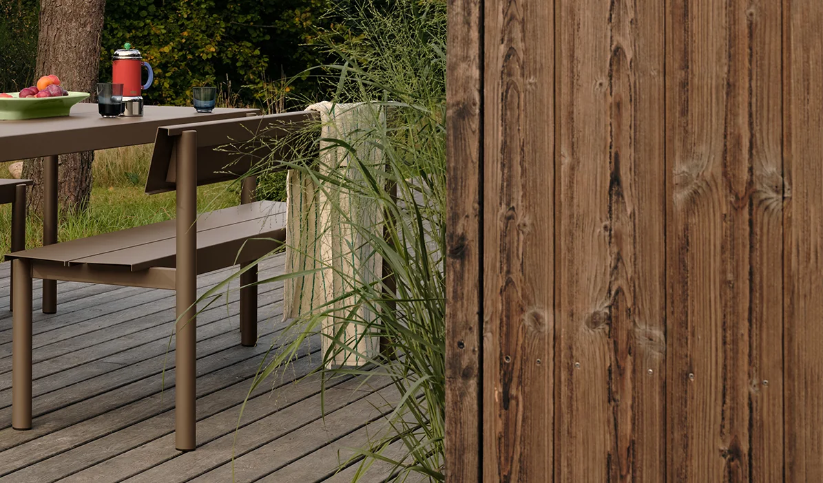 Brunt metall utendørsbord og benk på en treterrasse med frukt og drikke, ved siden av høyt gress og et tregjerde.