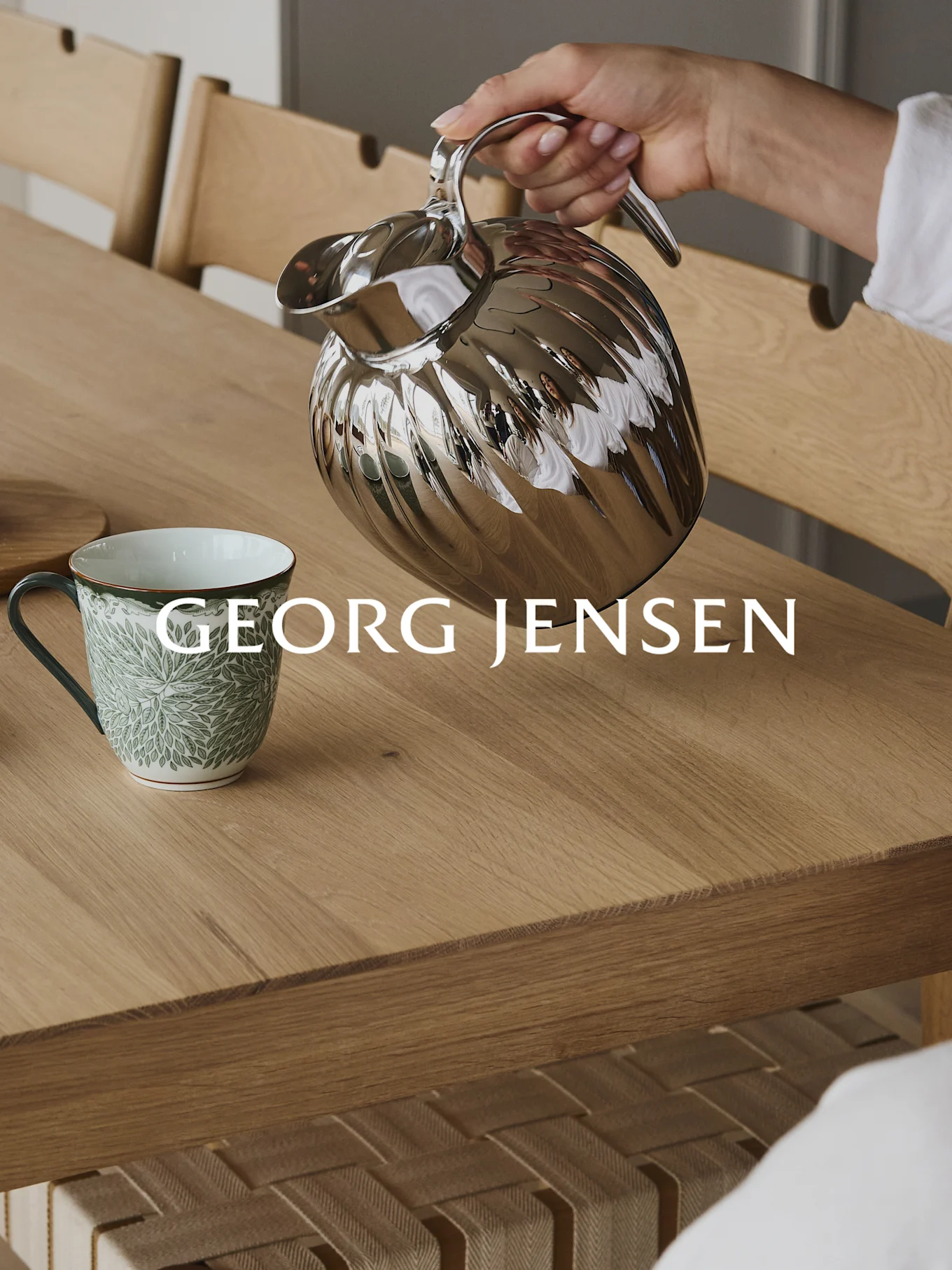 Hånd heller fra en sølvfarget riflet termokanne i et grønt mønstret krus på et trebord.
