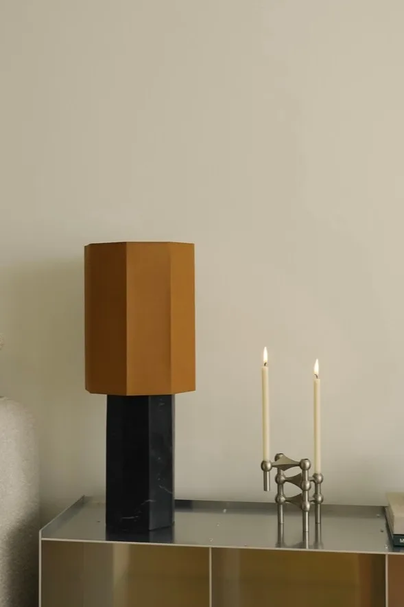 Moderne bordlampe med sekskantet brun skjerm og svart marmorbase, ved siden av en sølvfarget modulær lysestake med to tente hvite lys.