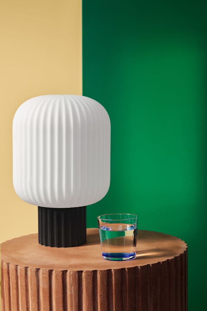Bordlampe i riflet glass fra Broste Copenhagen mot tofarget vegg i grønt og gult. 