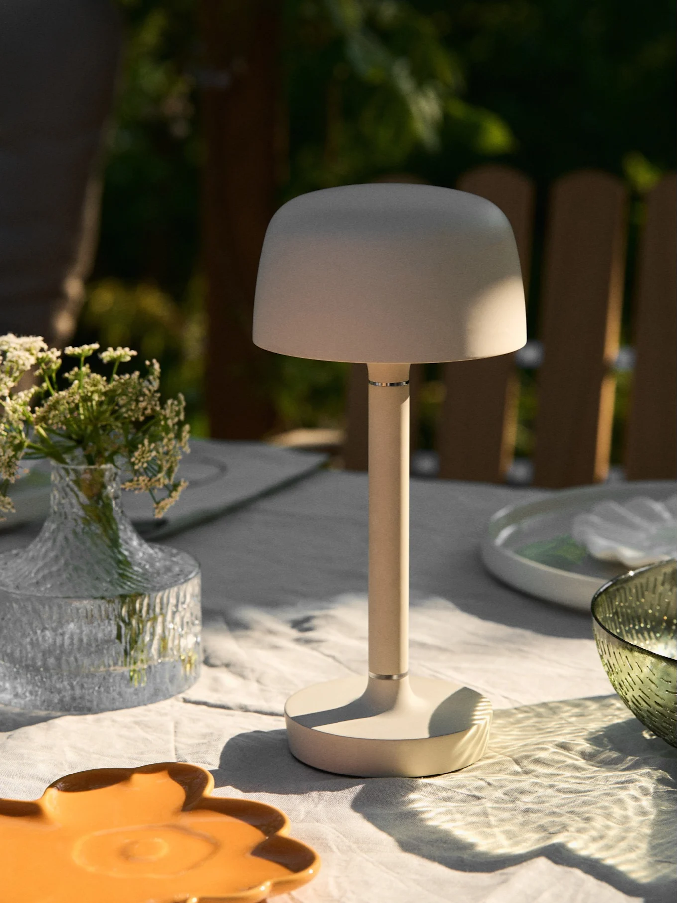 Beige bærbar bordlampe på et utebord med blomstervase og flekkete sollys.