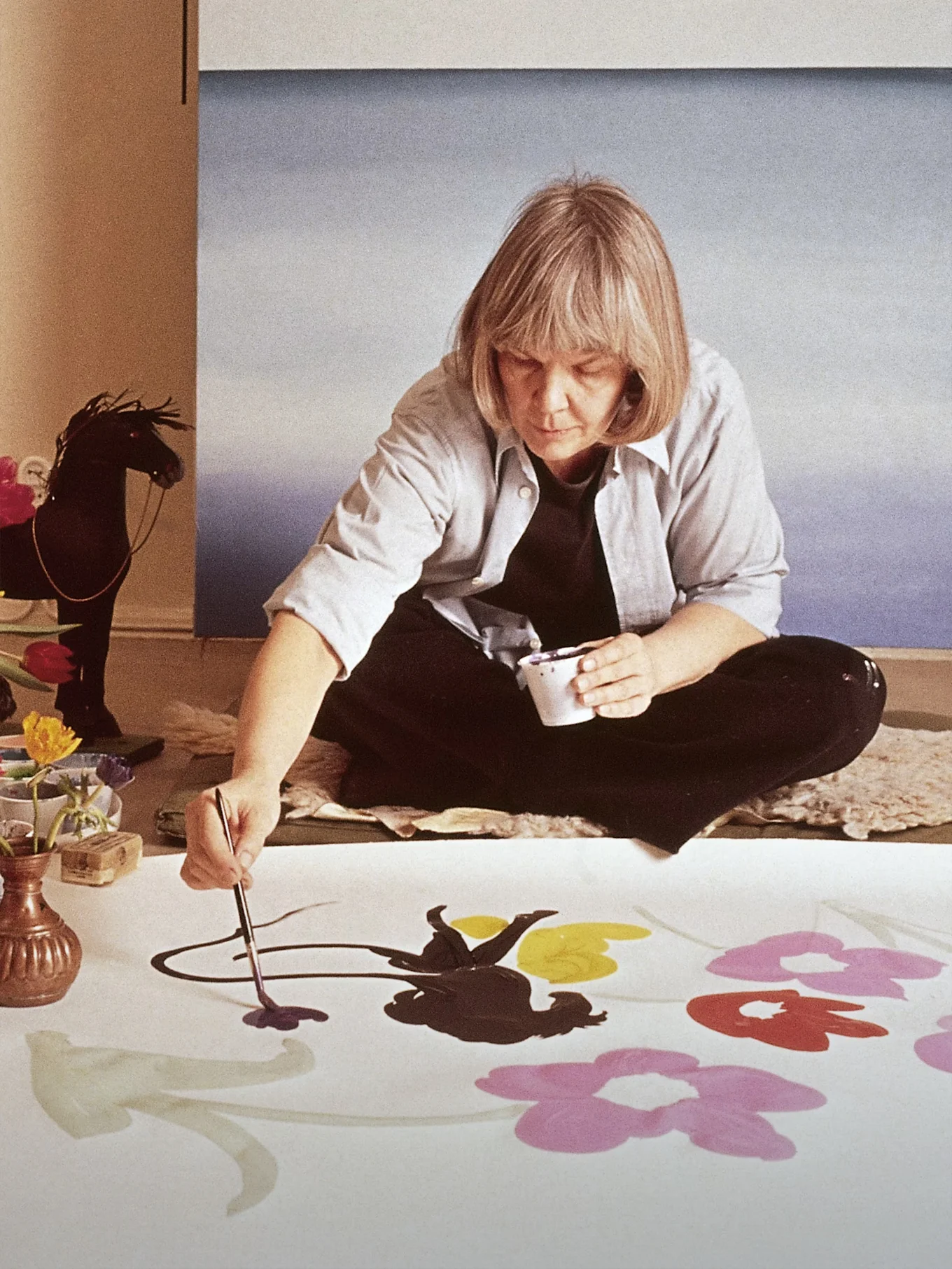 Kunstneren Marimekko Maija Isola sitter på gulvet og maler store, fargerike blomster på et hvitt stoff.