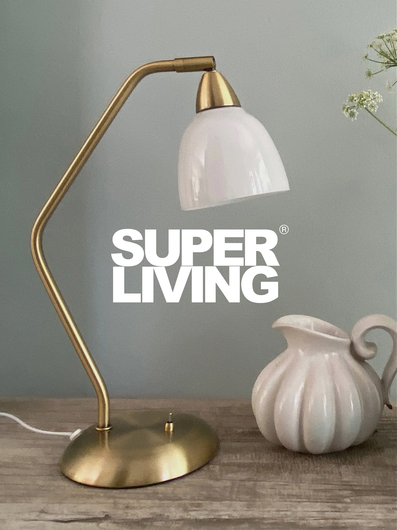 SUPER LIVING-merkevare over en messingbordlampe med hvit skjerm og en hvit, riflet mugge på et trebord.