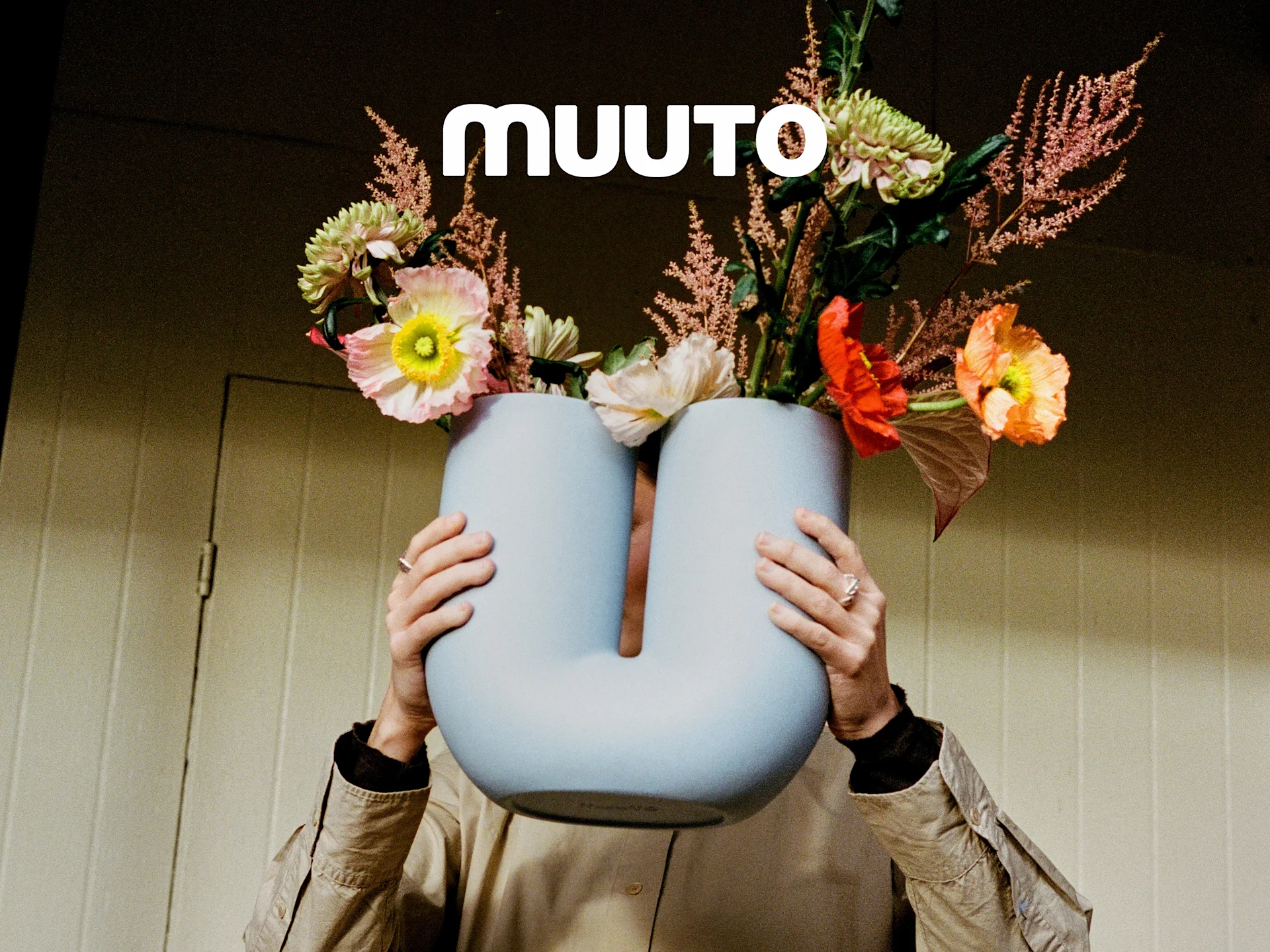 Person holder en lyseblå, U-formet vase med en livlig bukett rosa, oransje og grønne blomster. Muuto-logo.