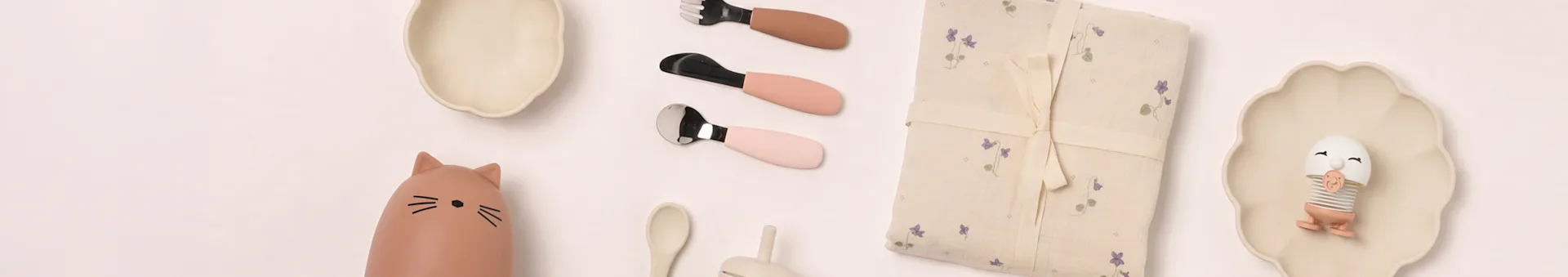 Babyutstyr flat lay: beige tallerkener, katteflaske, rosa bestikk, blomstret teppe, eggleketøy.