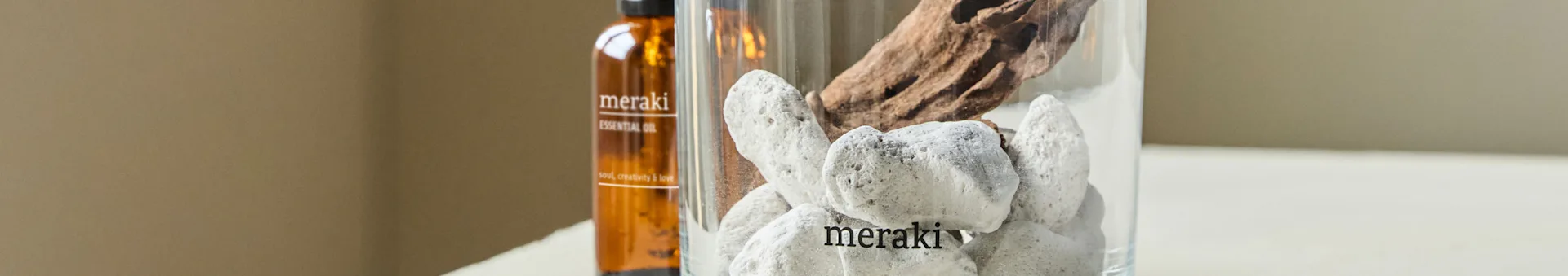 En brun flaske med eterisk olje og en klar vase med hvite steiner og drivved. Flasken og vasen er begge merket 'meraki'.
