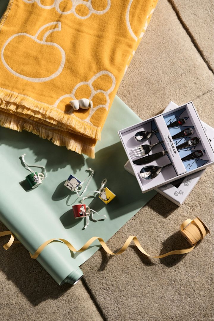 Kreative gaveideer til babyshower, her ser du en samling av mulige babyshowergaver. Det er minikrus, mummifigurer, et gult teppe og et sett med barnebestikk.