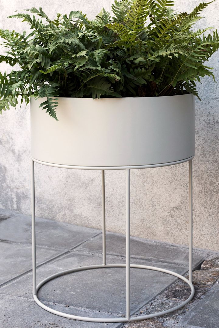 Den store runde Plant Box i metall fra Ferm Living med en stor grønn bregne, står på en uteplass.